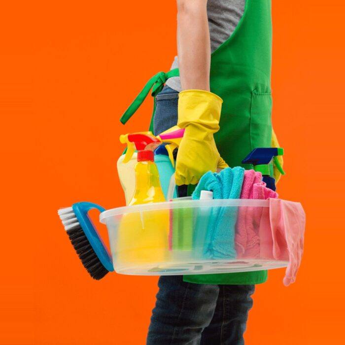 Person in a green apron and yellow rubber gloves carrying a caddy of cleaning supplies against an orange background
