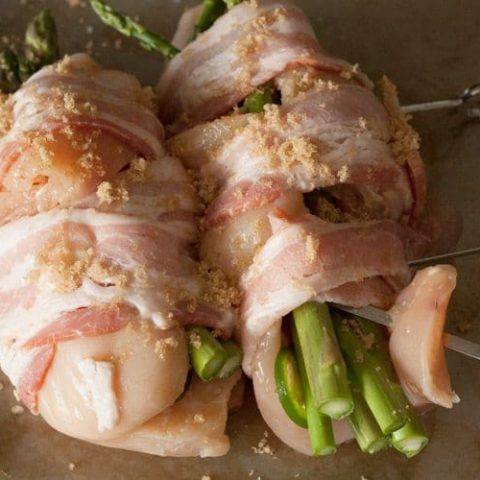 Two chicken breasts stuffed with asparagus and jalapeño, wrapped in bacon, sprinkled with brown sugar and threaded on metal skewers, ready for the grill