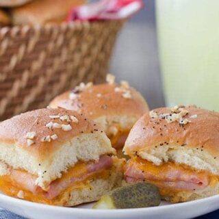 Two ham and cheddar potluck sandwiches on a white plate with a pickle and a glass of lemonade in the background