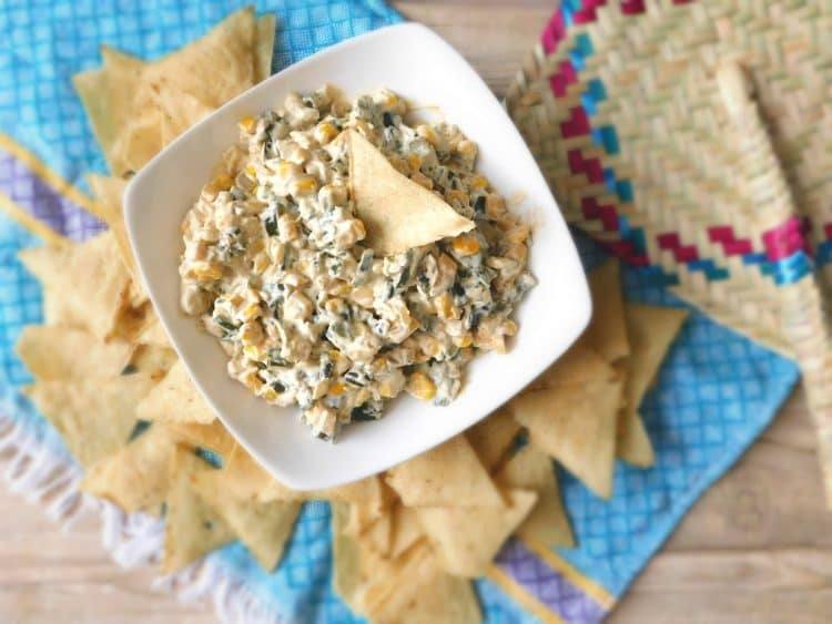 Overhead view of roasted poblano pepper dip in a white bowl surrounded by tortilla chips on a turquoise woven cloth