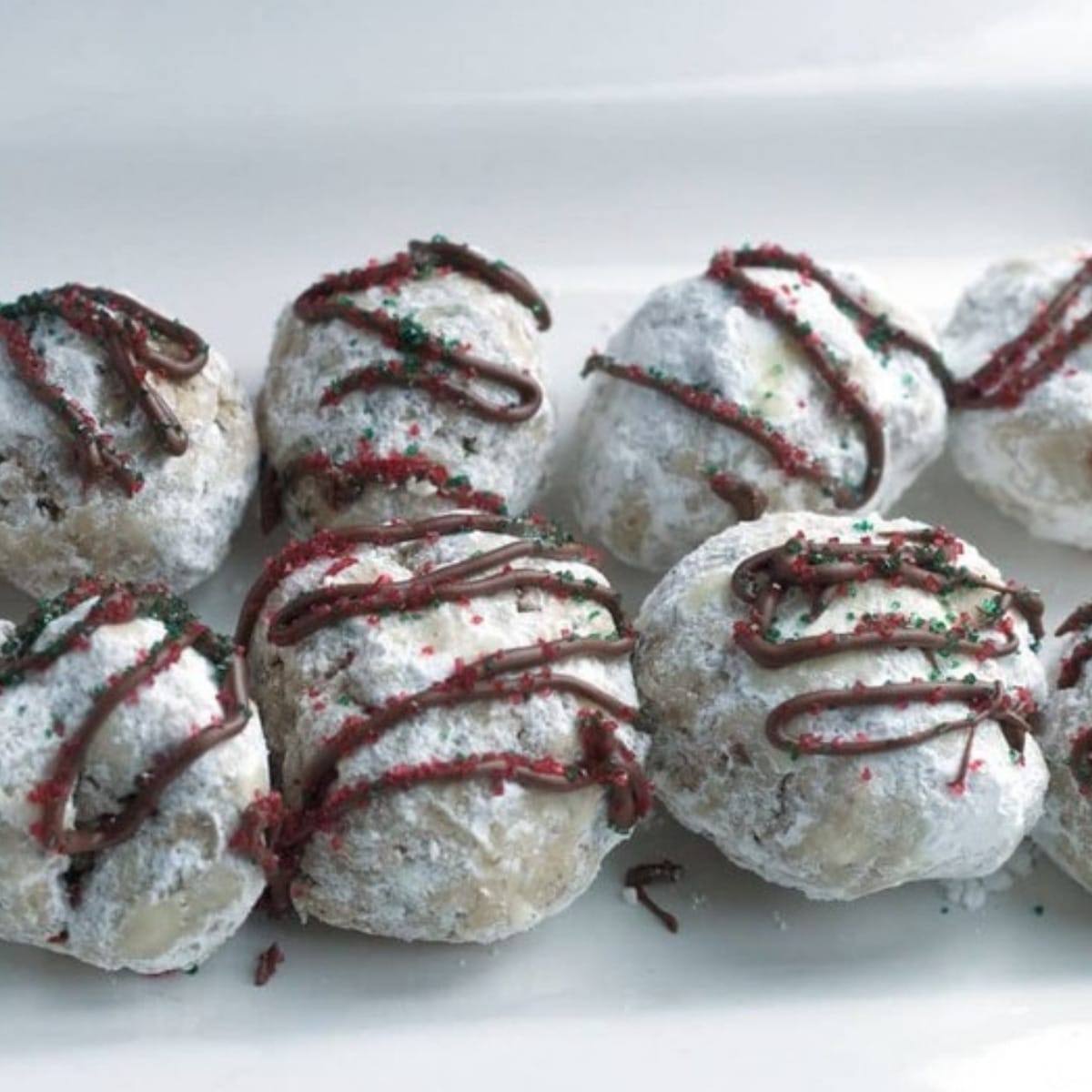 Peppermint snowball cookies dusted in powdered sugar with a chocolate drizzle and red and green sprinkles
