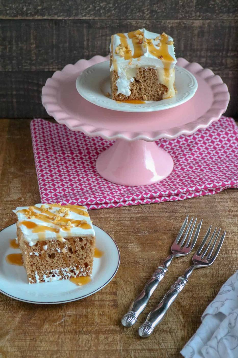Apple spice poke cake slice topped with whipped topping, caramel and chopped nuts on a plate beside a pink cake stand and two forks