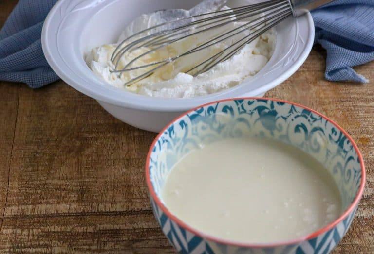 Thick heavy whipping cream in a white bowl with a whisk and sweetened condensed milk in a teal patterned bowl on a wood table next to a blue kitchen towel