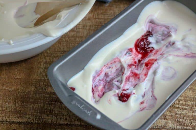 White bowl with No-Churn Ice Cream and wooden spoon next to a silver pan half-filled with No-Churn Ice Cream with berry pie filling swirled on top