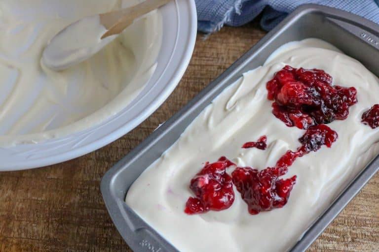 White bowl with wooden spoon in it next to a silver pan filled with No-Churn Ice Cream with berry pie filling swirled on top