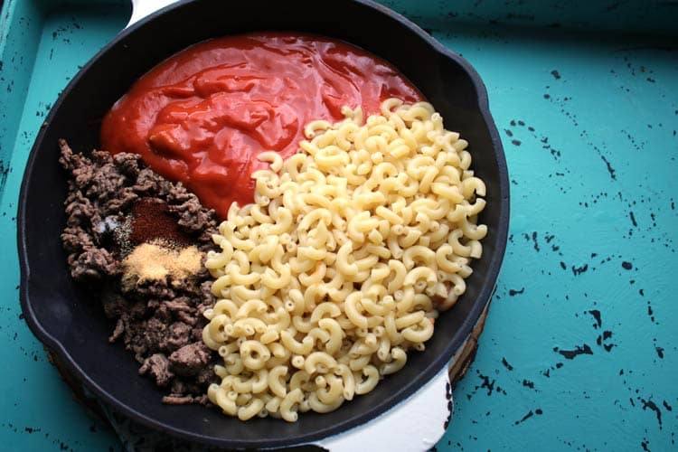 Chili mac ingredients in a cast iron skillet: browned ground beef, spices, tomato soup and cooked elbow macaroni