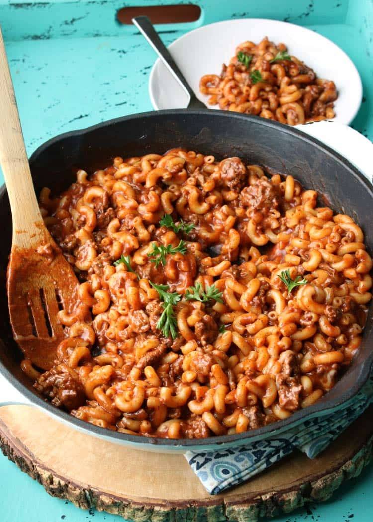Chili mac in a cast iron skillet with a wooden spoon and a plate of chili mac on the side