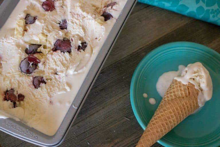 top-down view of the container filled with the finished black cherry white chocolate no-churn ice cream next to a single scoop on a sugar cone sitting on top of a blue plate