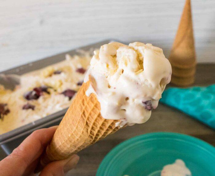 hand holding a scoop of black cherry white chocolate chunk no-churn ice cream on a sugar cone in front of a green dessert plate, cloth napkin and loaf pan containing the completed recipe