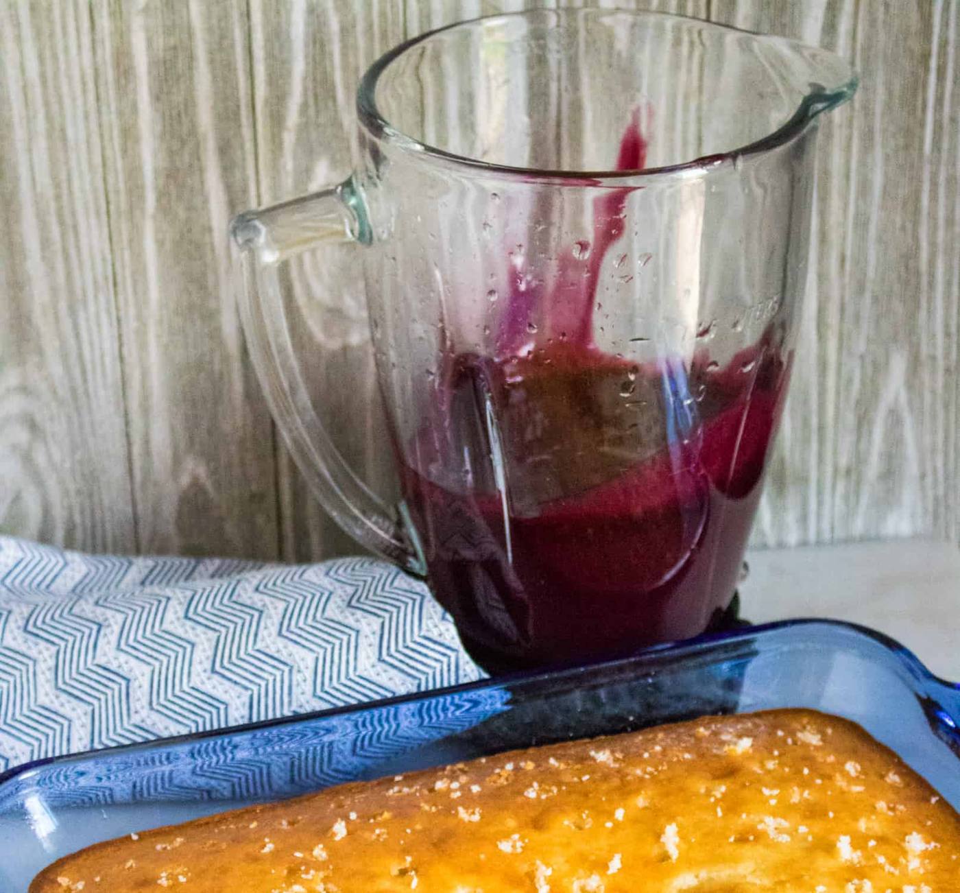 blueberry pie filling in a blender pitcher sitting behind a white sheet cake with holes poked in the top next to a blue striped cloth napkin
