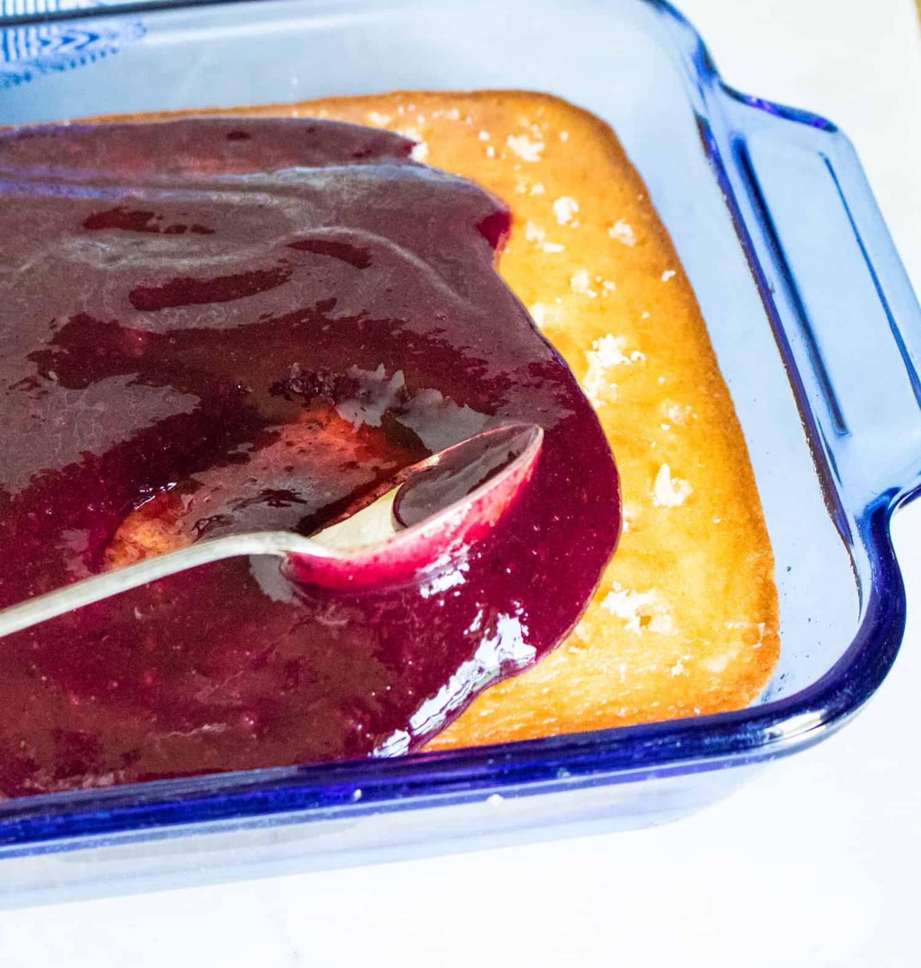 blueberry pie filling being spread on the top of the white sheet cake with a silver spoon