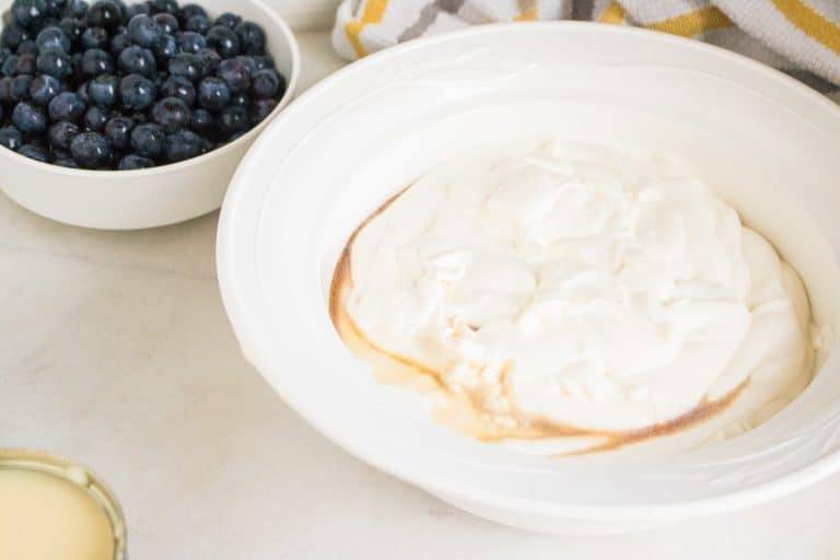 white ceramic bowl full of fresh blueberries in background and larger white ceramic bowl in foreground full of whipped heavy whipping cream