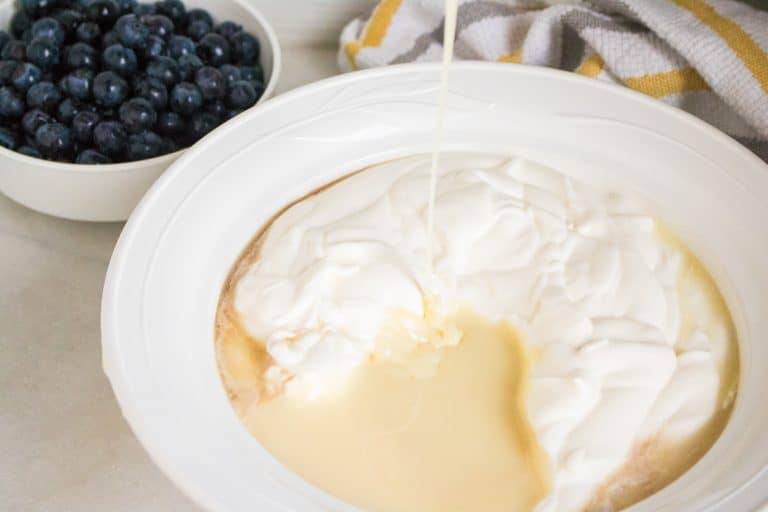 white ceramic bowl full of fresh blueberries in background and larger white ceramic bowl in foreground full of whipped heavy whipping cream for blueberry lemon no churn ice cream with sweetened condensed milk being poured into the whipping cream and yellow and white striped towel behind