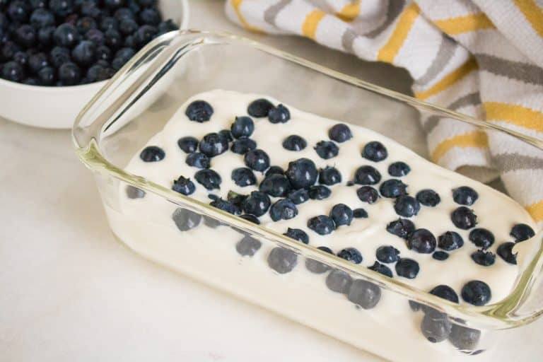 white ceramic bowl of fresh blueberries and yellow white and gray striped towel in background glass freezer safe container in foreground filled with cream mixture and topped with fresh blueberries for blueberry lemon no churn ice cream