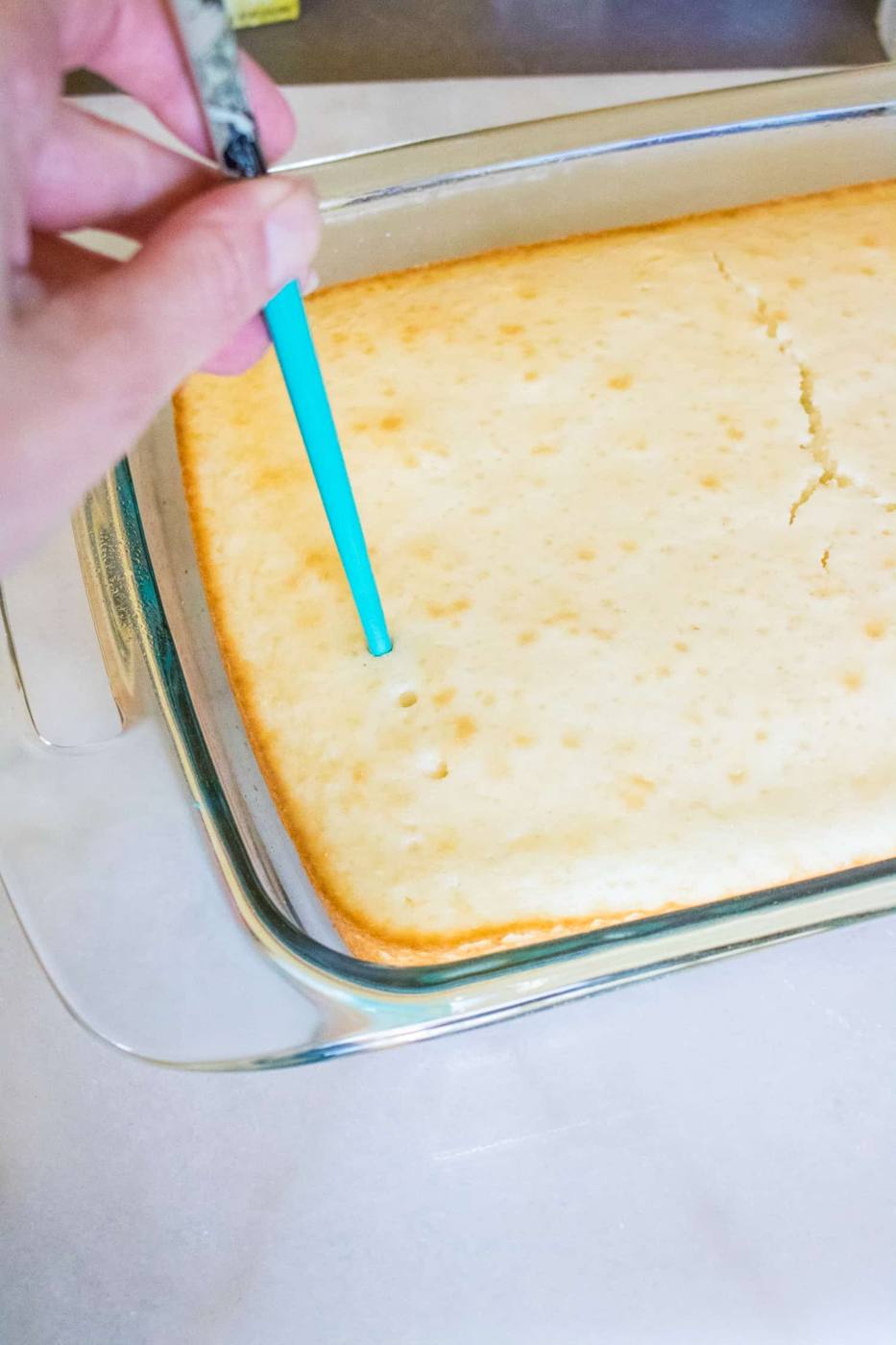 baked white cake for boozy pineapple pina colada poke cake in pan being poked with blue skewer