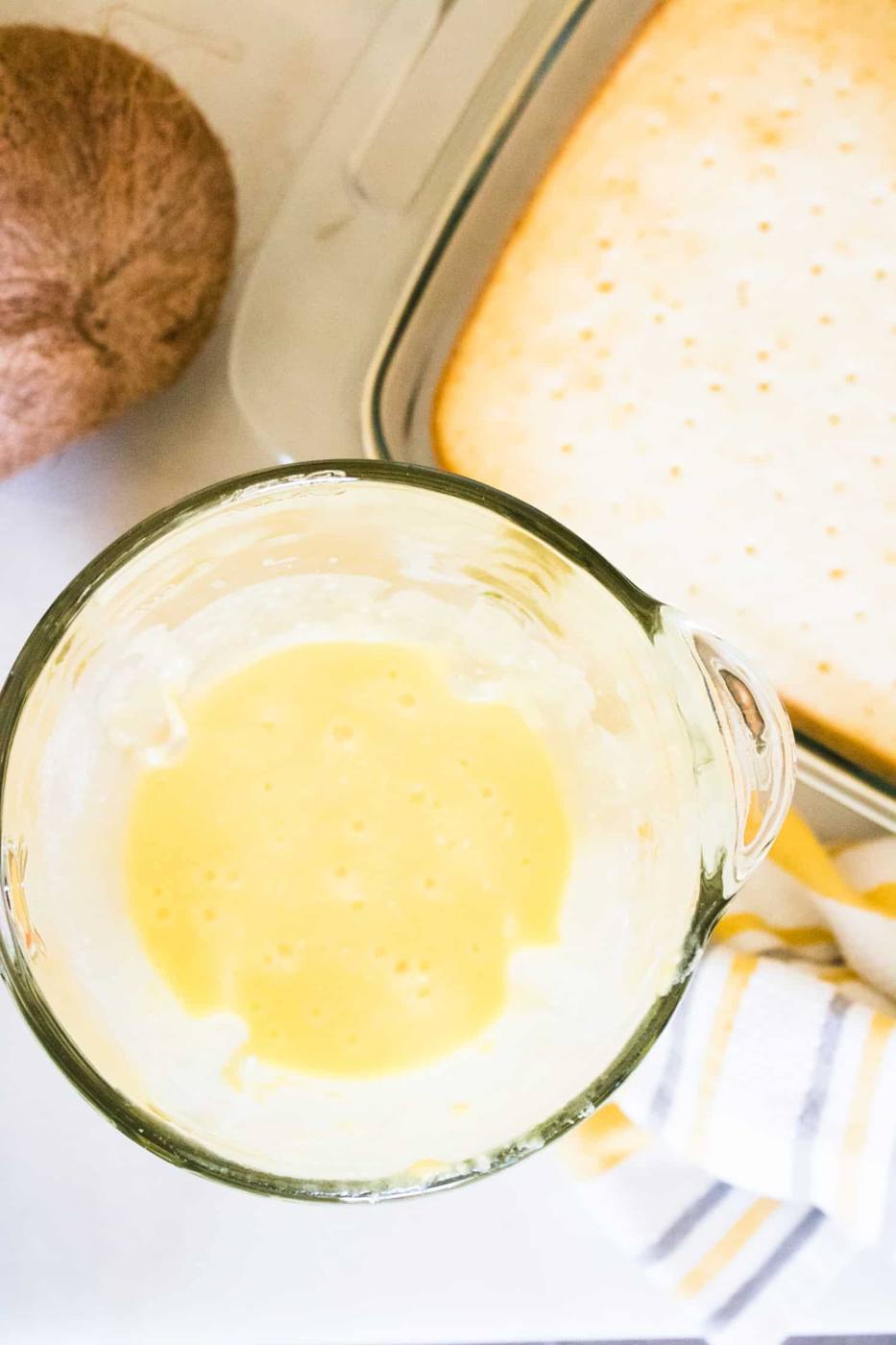 yellow boozy pineapple pina colada poke cake filling in glass measuring cup sitting next to white cake in pan