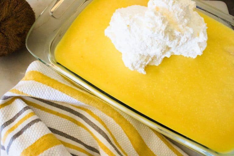 clear glass pan with yellow boozy pineapple pina colada poke cake with large white dollop of whip topping and white brown and gold striped towel in foreground