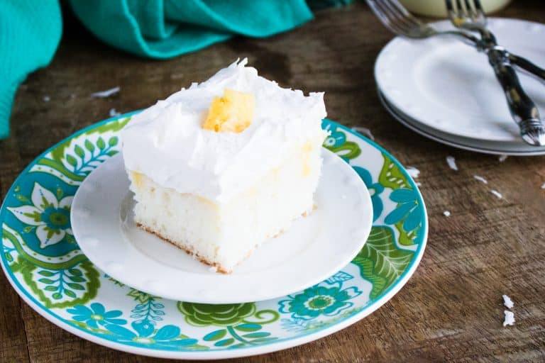 white boozy pineapple pina colada poke cake sitting on white plate on a blue and green patterned liner plate and white plate with silver forks in background