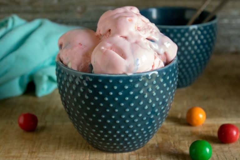 blue polka dot ceramic bowl with 3 scoops of pink bubble gum no-churn ice cream in it, blue towel in background and red orange and green gumballs scattered about