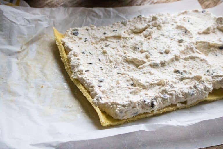 cannoli cream cake unrolled laying on parchment paper with cannoli cream filling spread on top