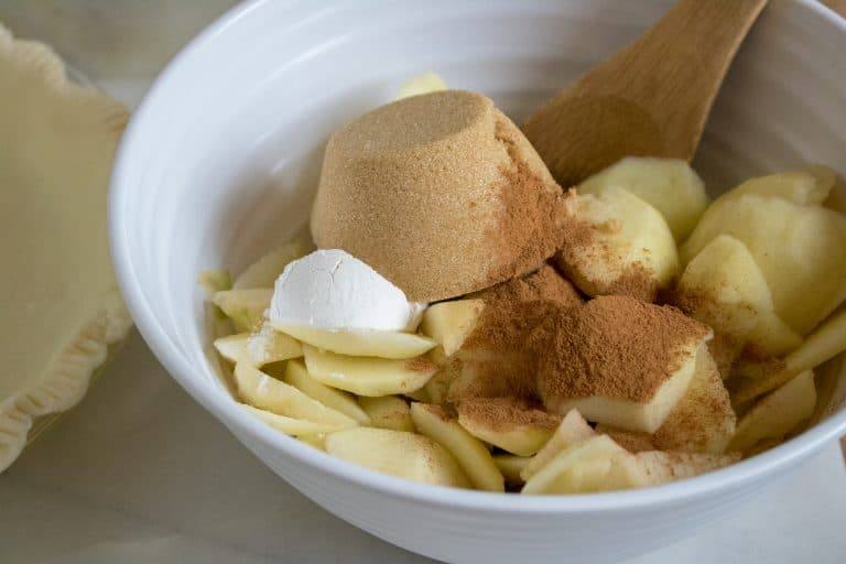 white bowl with sliced apples, brown sugar, cinnamon for Caramel Apple Crumble Pie being stirred by a wooden spoon