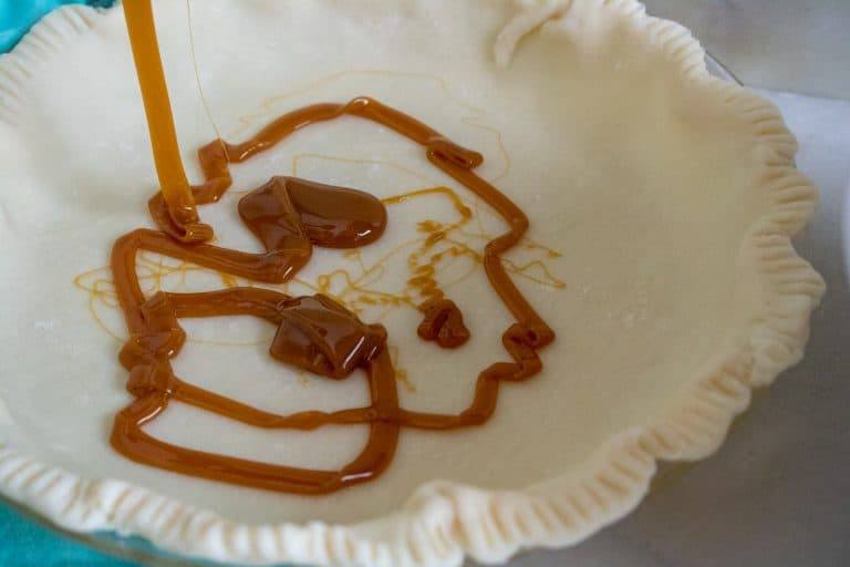 Pie plate with uncooked pie crust in it for Caramel Apple Crumble Pie being drizzled with caramel sauce