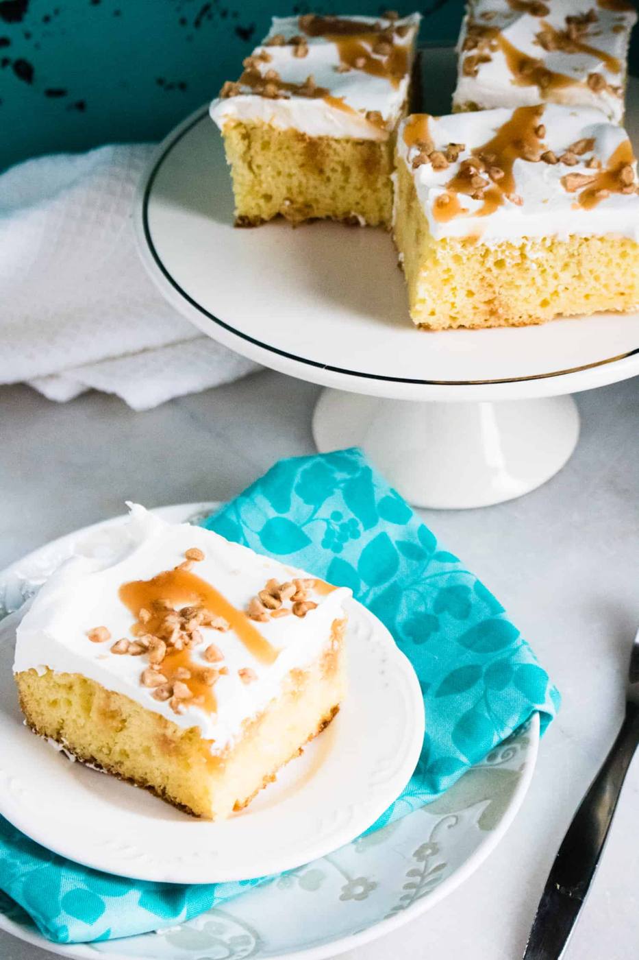top-down view of Caramel Toffee Poke Cake on a white cake stand behind a single slice served on a white plate on top of a blue print napkin