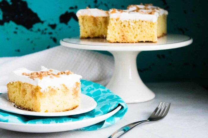 Caramel Toffee Poke Cake on a white cake stand next to a slice served on a blue dessert plate