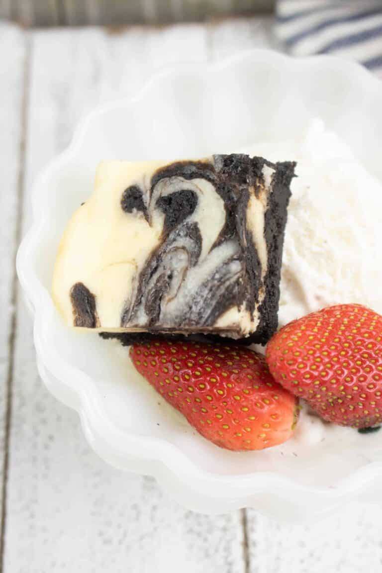 One serving of Cheesecake Brownies served in a white scalloped bowl with strawberries and whipped cream
