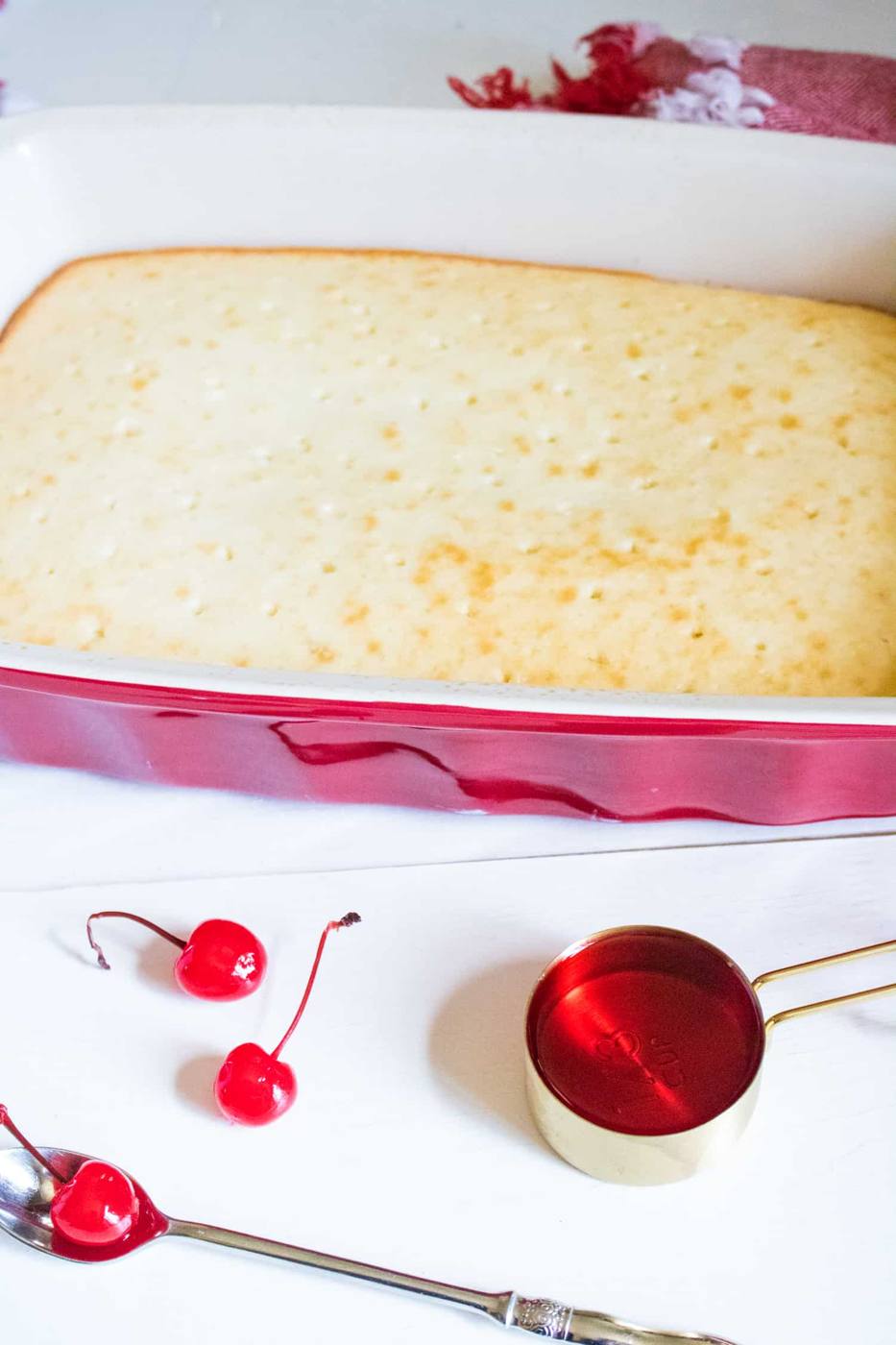 Baked white cake with holes poked on the top sitting next to a measuring cup full of cherry syrup