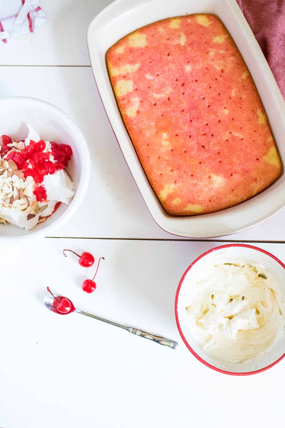 Baked cake with cherry syrup soaking into the top sitting next to a bowl of whipped cream almond topping