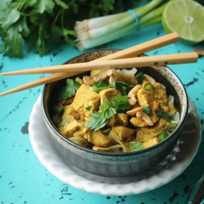 Chicken curry with noodles in a dark bowl topped with cashews and herbs, with chopsticks