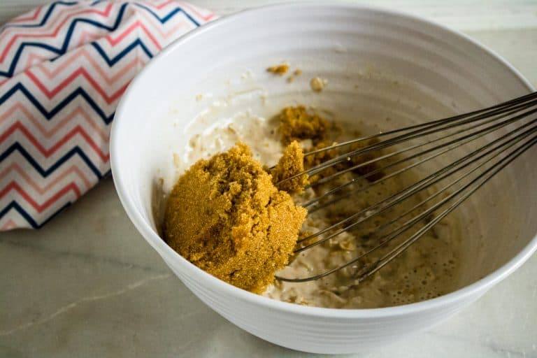 brown sugar being mixed into oat mixture with silver whisk for Chocolate Chocolate Chip Oatmeal Muffins in white bowl with zig zag napkin in background