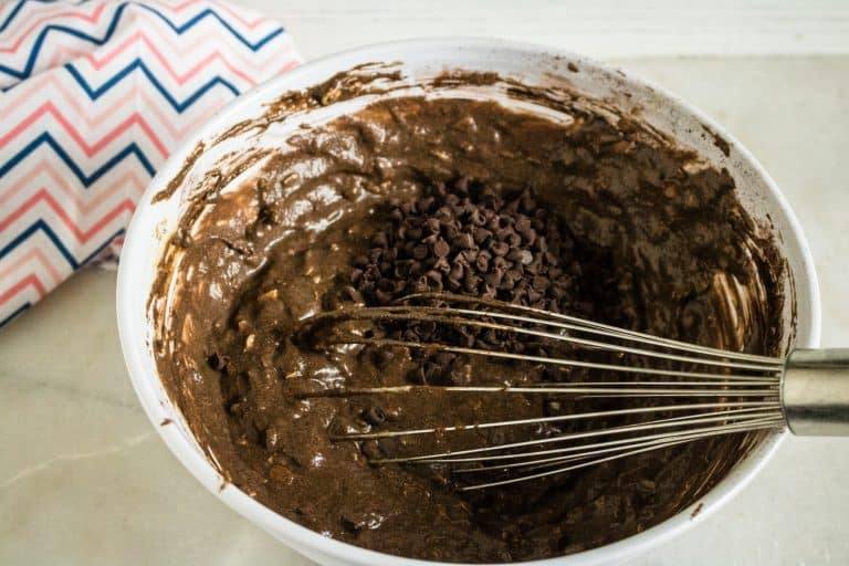 mini chocolate chips being mixed into batter for Chocolate Chocolate Chip Oatmeal Muffins with silver whisk with zig zag print napkin in background