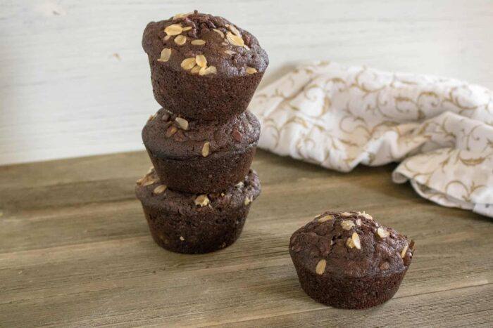 three stacked chocolate chocolate chip oatmeal muffins next to one muffin on a wood cutting board in front of a white print kitchen towel