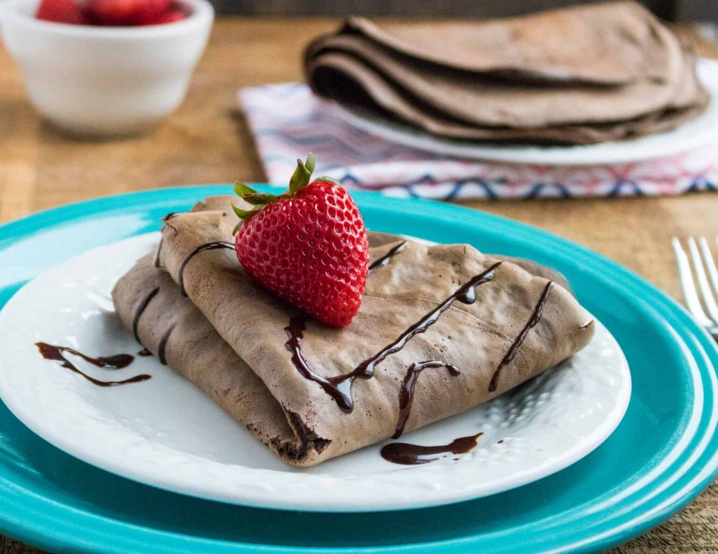 chocolate dessert crepes topped with a strawberry and chocolate sauce, served on a white plate placed on top of a blue plate