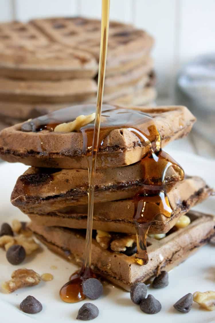 chocolate waffles on a white plate with syrup being poured over them.
