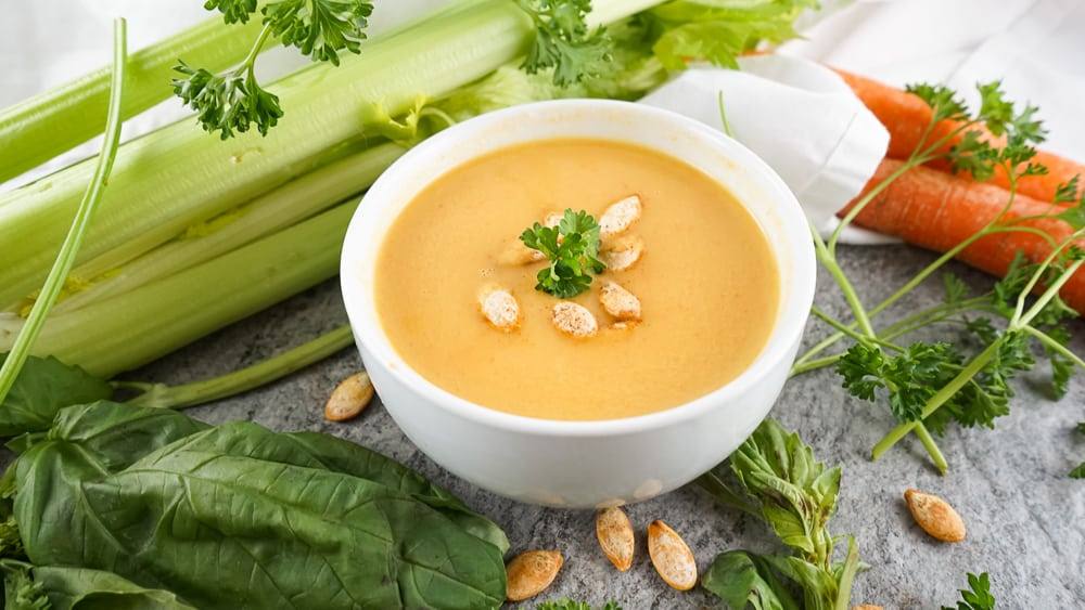 Bowl of butternut squash soup on a gray table with celery, carrots, parsley, and pumpkin seeds