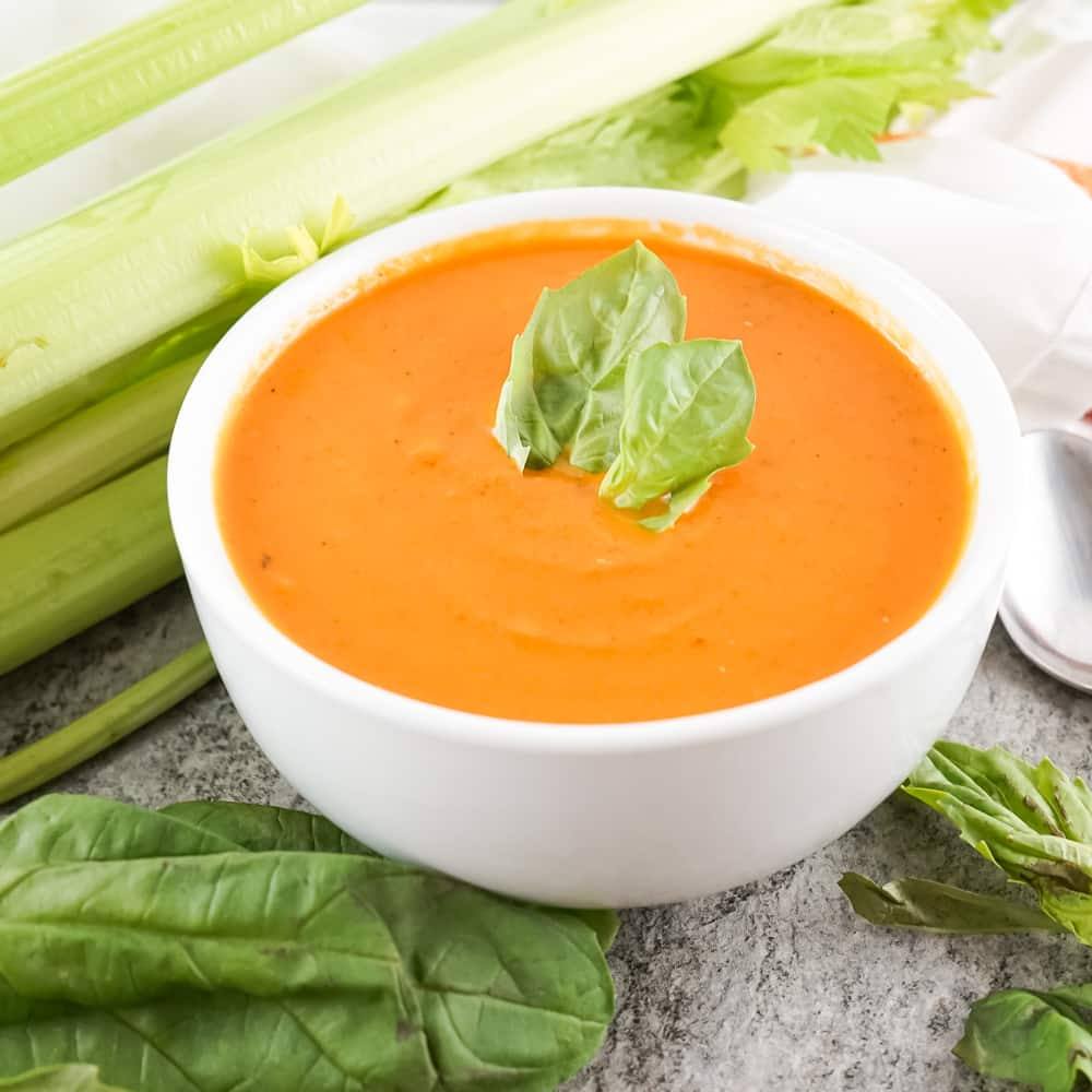 Creamy tomato soup in a white bowl with basil leaves, celery and carrots