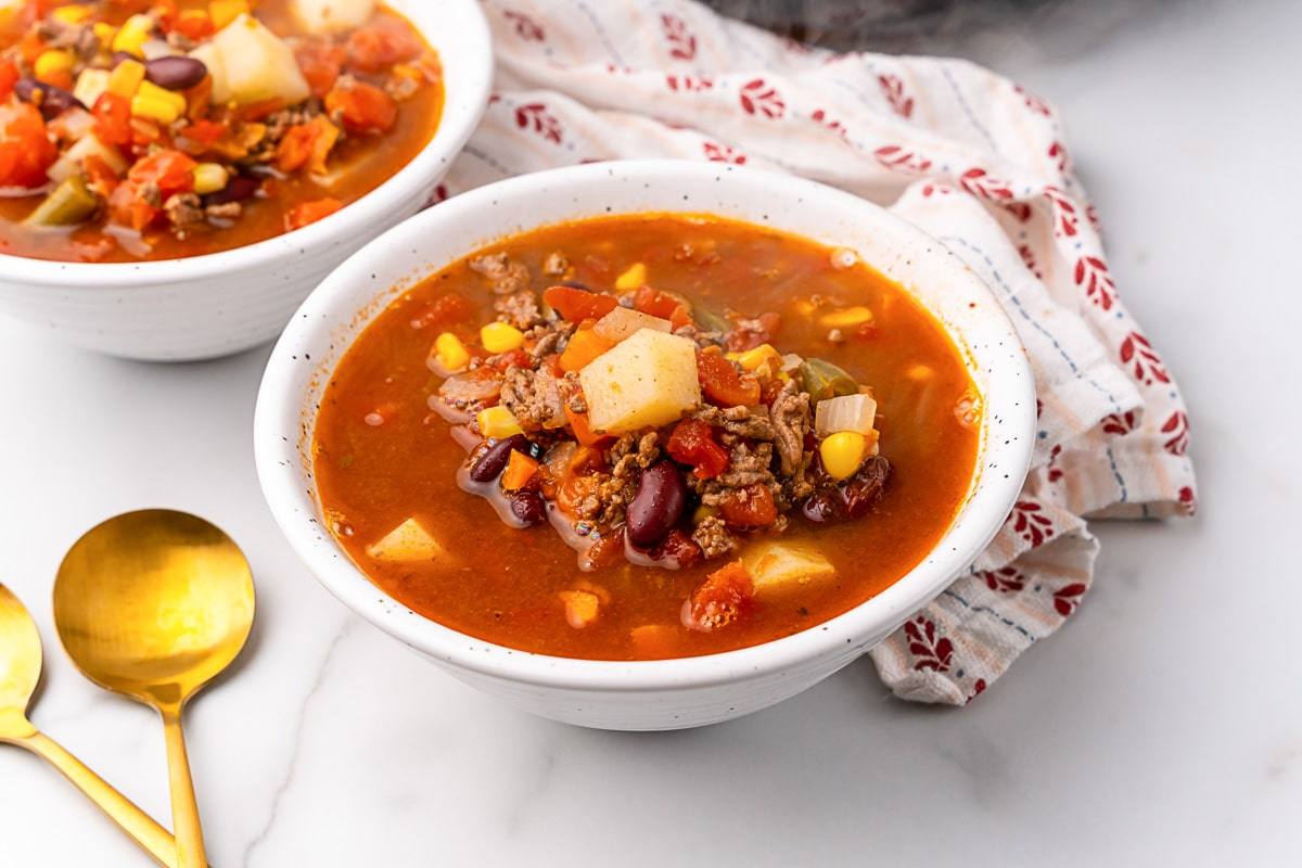 Angled view of Crazy-Easy Cowboy Soup in a speckled white bowl with gold spoons alongside