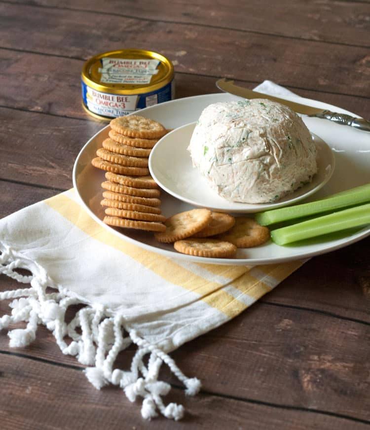 Tuna cheese ball on a white plate with stacked crackers and celery sticks on a striped towel
