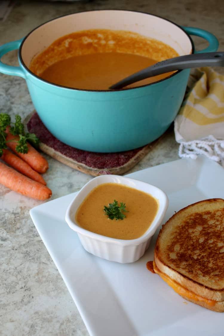 Creamy carrot soup with parsley in a white ramekin beside a grilled cheese sandwich, with a teal pot of soup behind it