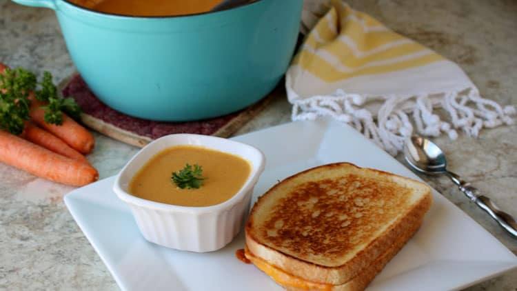soup in white dish next to sandwich on white plate with carrots, a blue pot and yellow and white towel in the background
