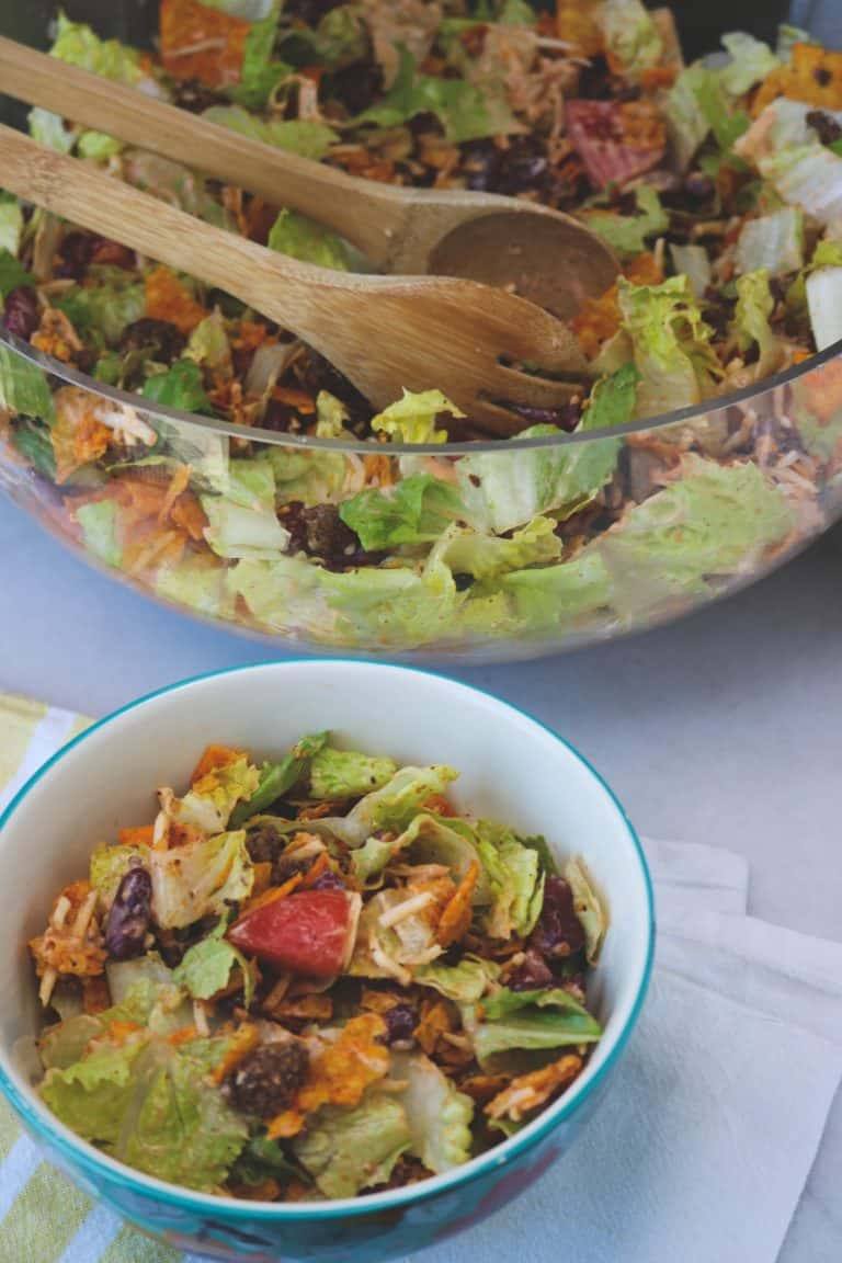 Doritos Taco Salad in a serving bowl sitting next to a large glass bowl filled with Doritos Taco Salad with wooden tongs