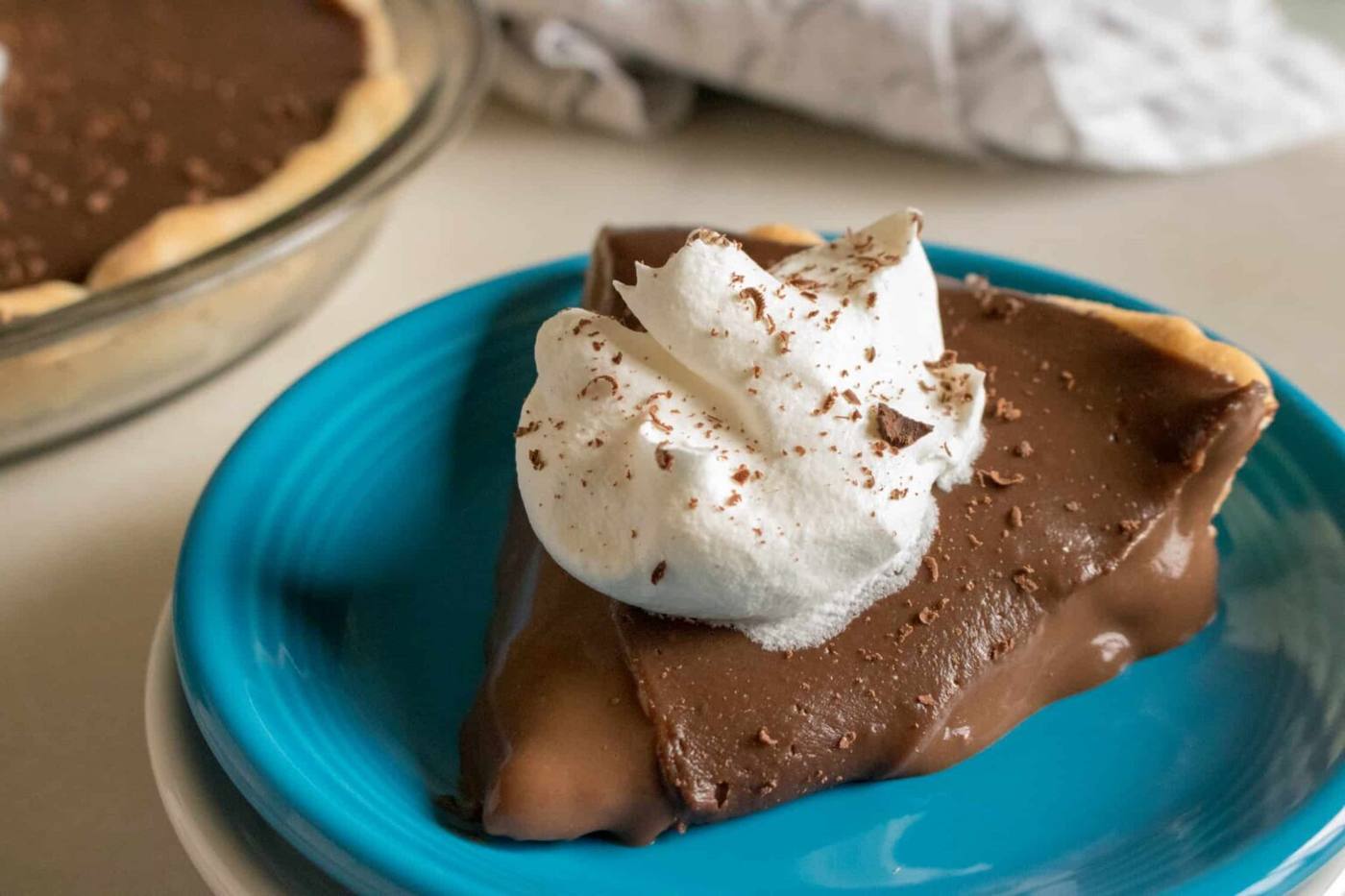 One slice of chocolate cream pie cut from the whole pie and served on a blue dessert plate and topped with whipped cream and shaved chocolate