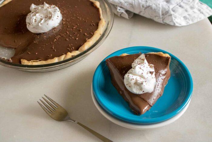 One slice of chocolate cream pie cut from the whole pie and served on a blue dessert plate and topped with whipped cream and shaved chocolate
