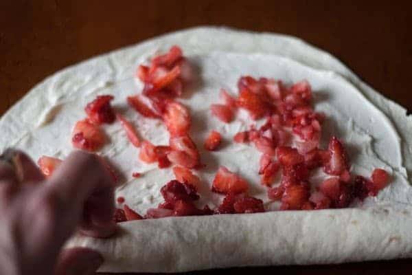 Hand rolling up a flour tortilla filled with cream cheese and chopped strawberries