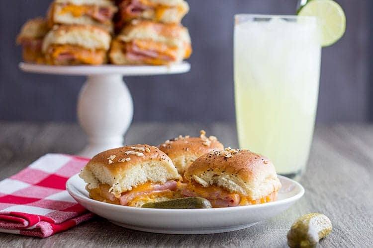 Ham and cheddar potluck sandwiches on a small white plate with a glass of lemonade and more sandwiches on a cake stand behind