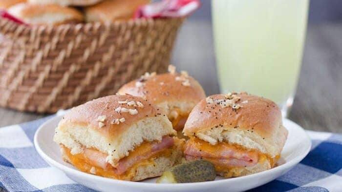 Three ham and cheddar potluck slider sandwiches on a plate with a pickle and a glass of lemonade
