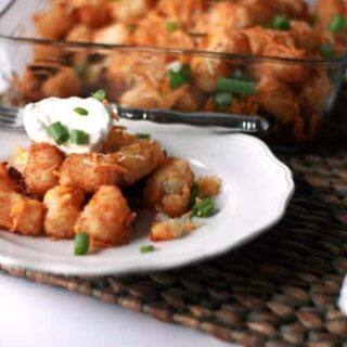 Plate of taco tater tot casserole topped with sour cream and green onions in front of the baking dish